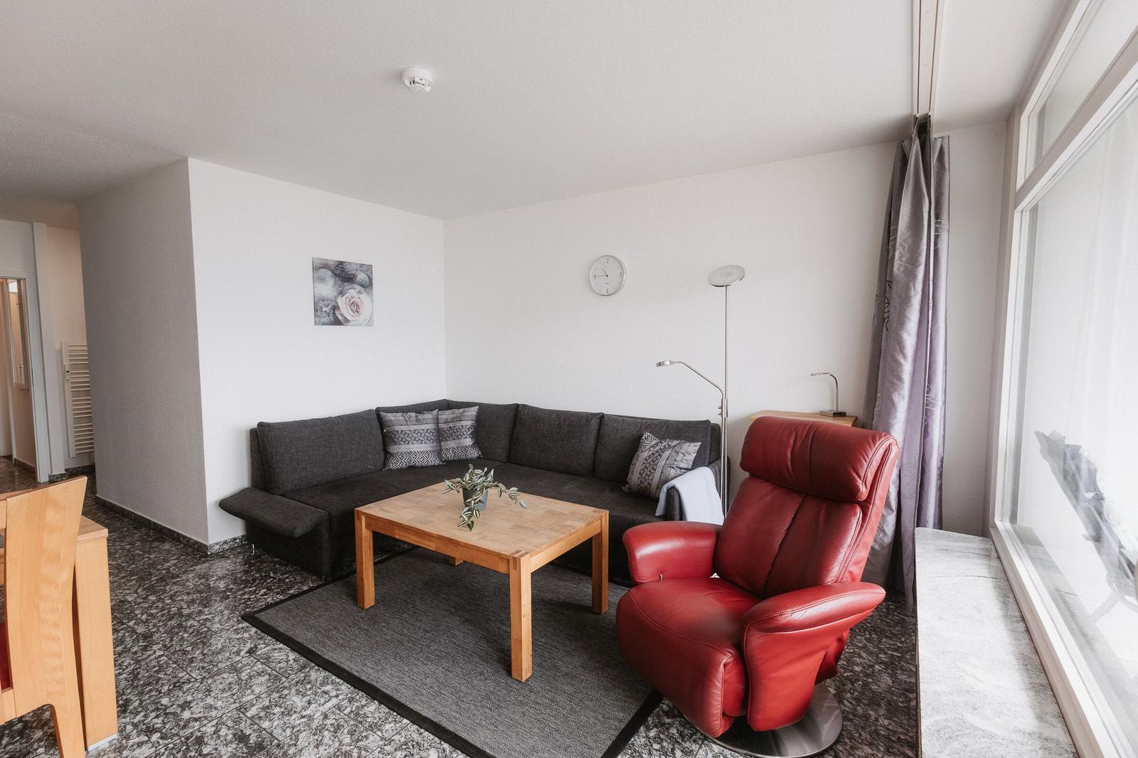 Wohnzimmer mit grauem Sofa, rotem Sessel und Holztisch. Große Fenster und Wanduhr sichtbar.