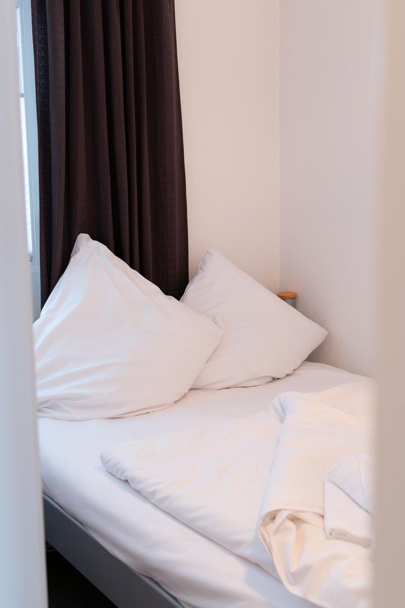 Ein Bett mit weißen Kissen und Bettwäsche. Dunkle Vorhänge sind am Fenster angebracht.