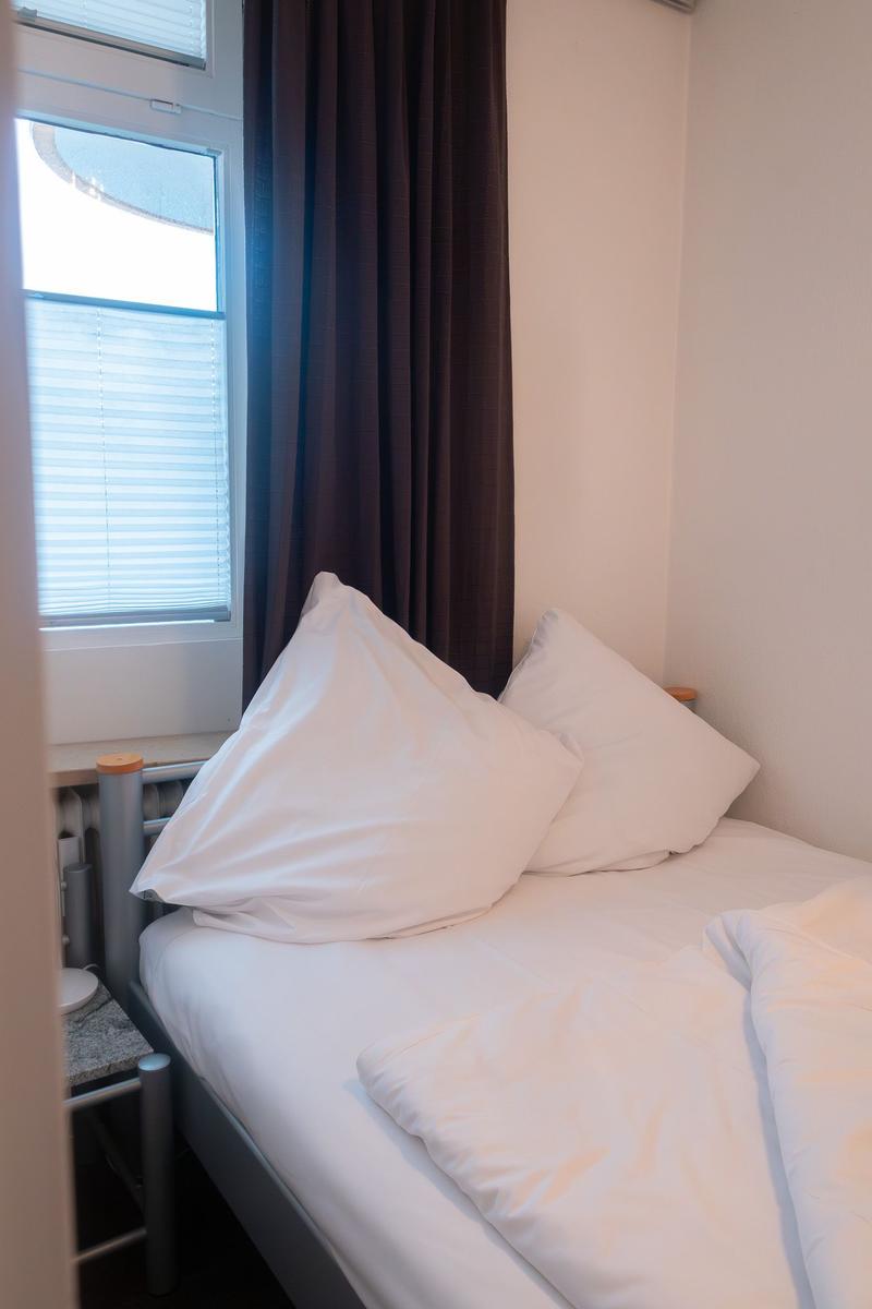 Ein Bett mit weißen Kissen und Bettwäsche neben einem Fenster mit Jalousie und dunklen Vorhängen.