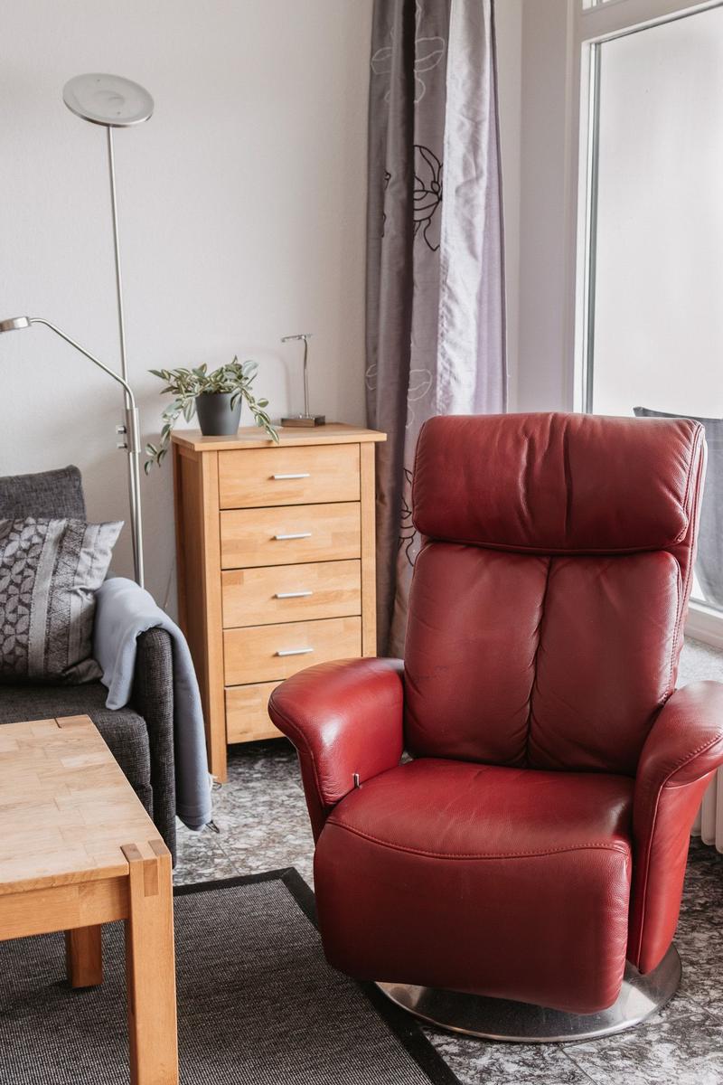 Ein roter Sessel steht vor einem Fenster. Neben ihm ein Holzschrank und eine Couch.