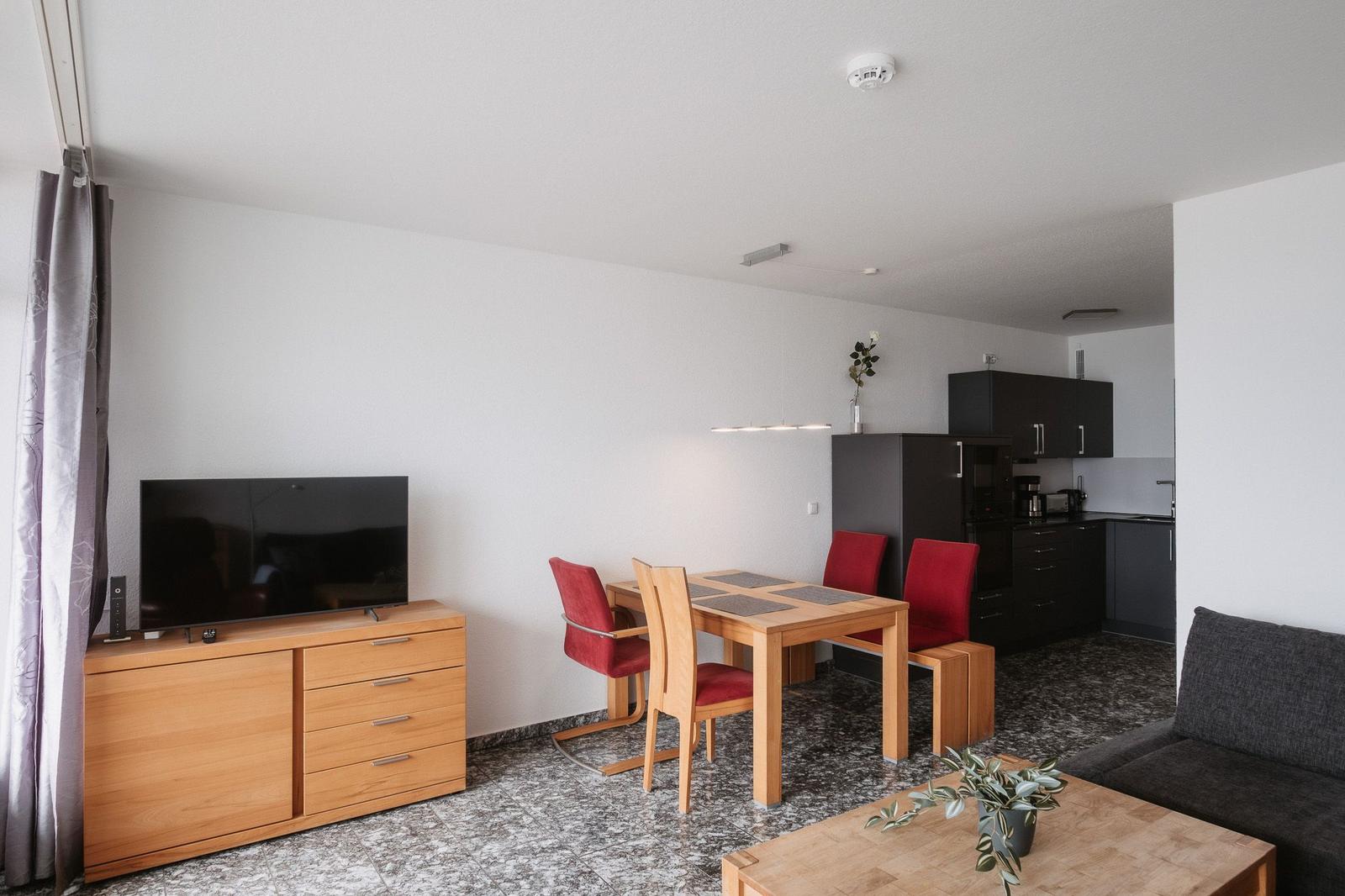 Wohn- und Essbereich mit Küchenzeile, TV und Sofa. Holztisch mit roten Stühlen.