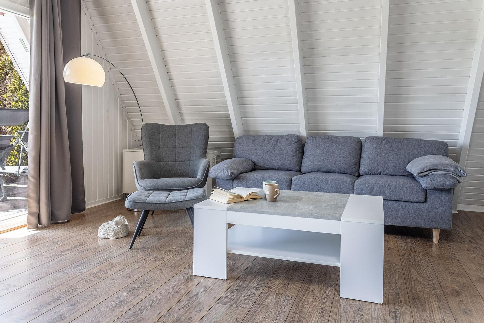 Wohnzimmer mit grauem Sofa, Sessel und weißem Couchtisch. Holzboden und weiße Schrägdecke.