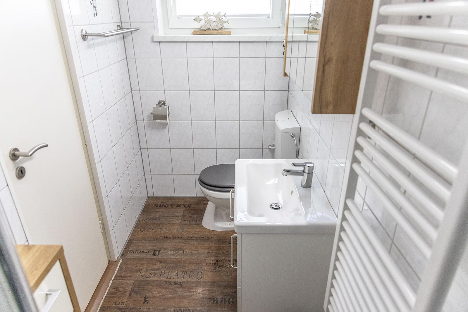 Badezimmer mit Toilette, Waschbecken und Dusche. Holzoptik Boden, weißer Fliesenwand.