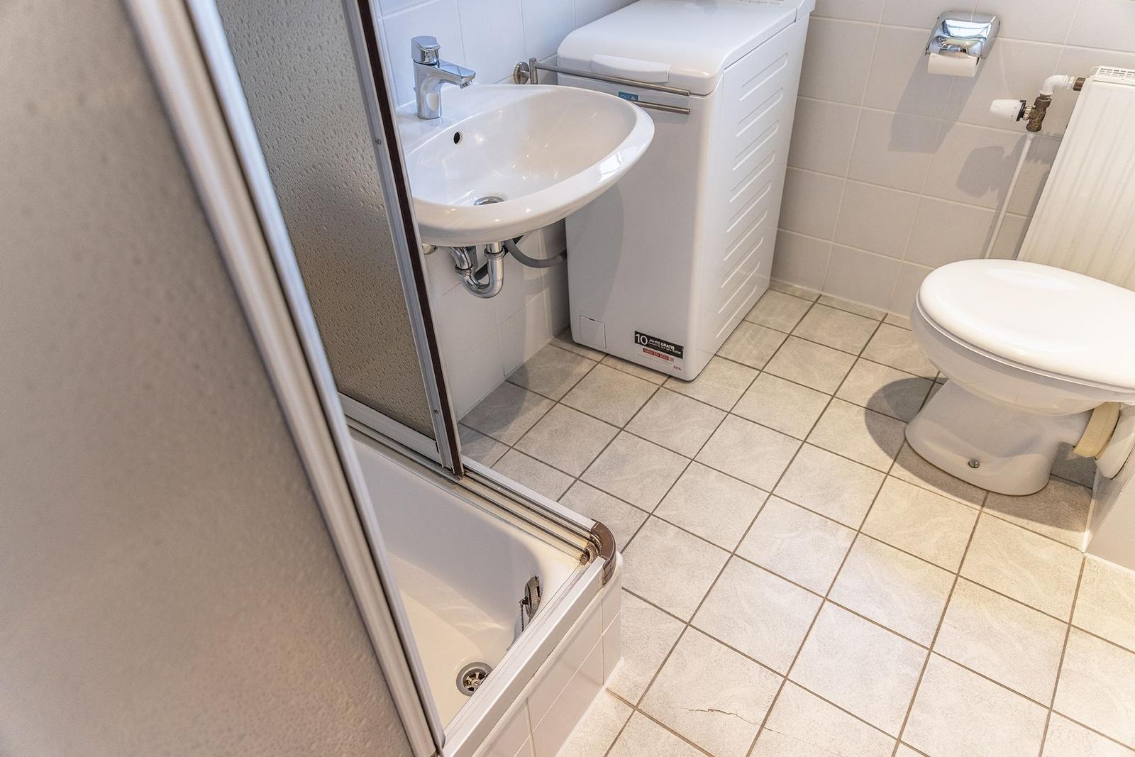 Badezimmer mit Dusche, Waschbecken und Toilette. Weiße Fliesen und ein Waschgerät.