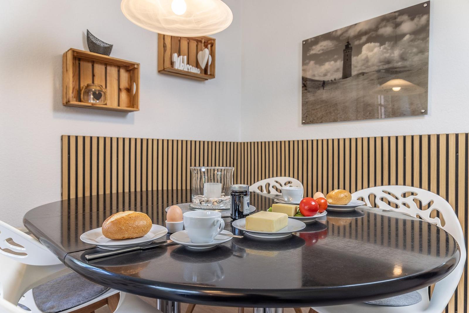 Runde Tisch mit Frühstück, weißen Stühlen und Wandbild mit Leuchte.