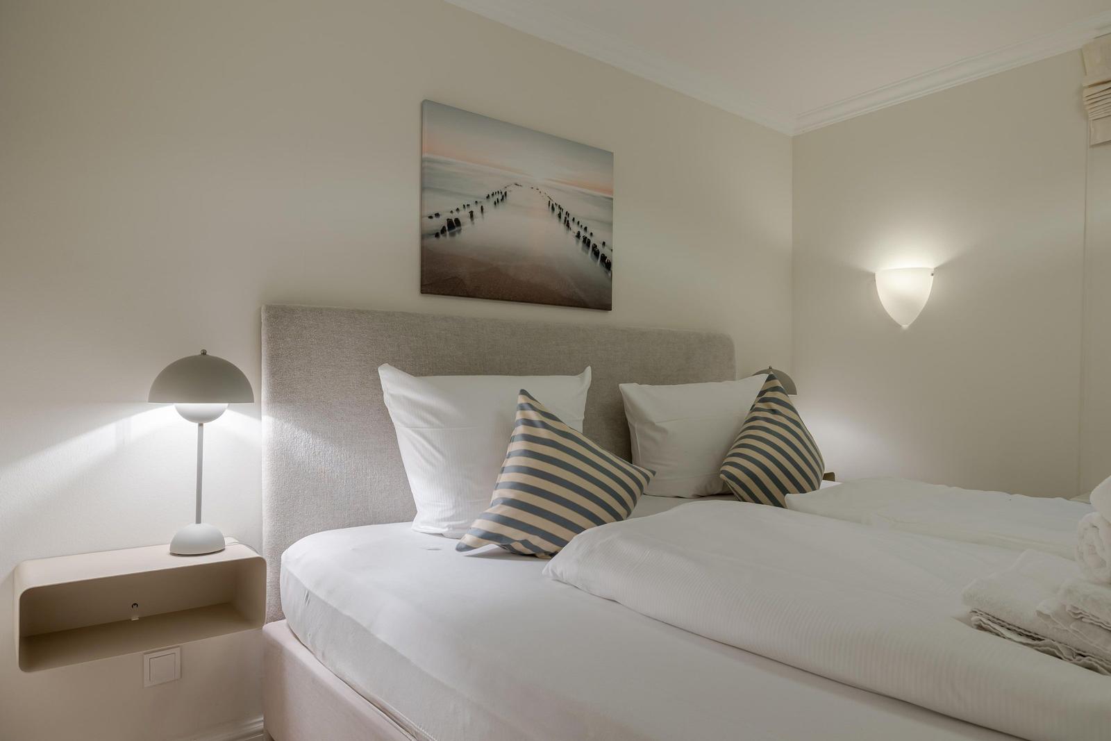 Schlafzimmer mit Bett, Kissen und Wandbild. Beleuchtung durch Stehlampe und Wandlampe.