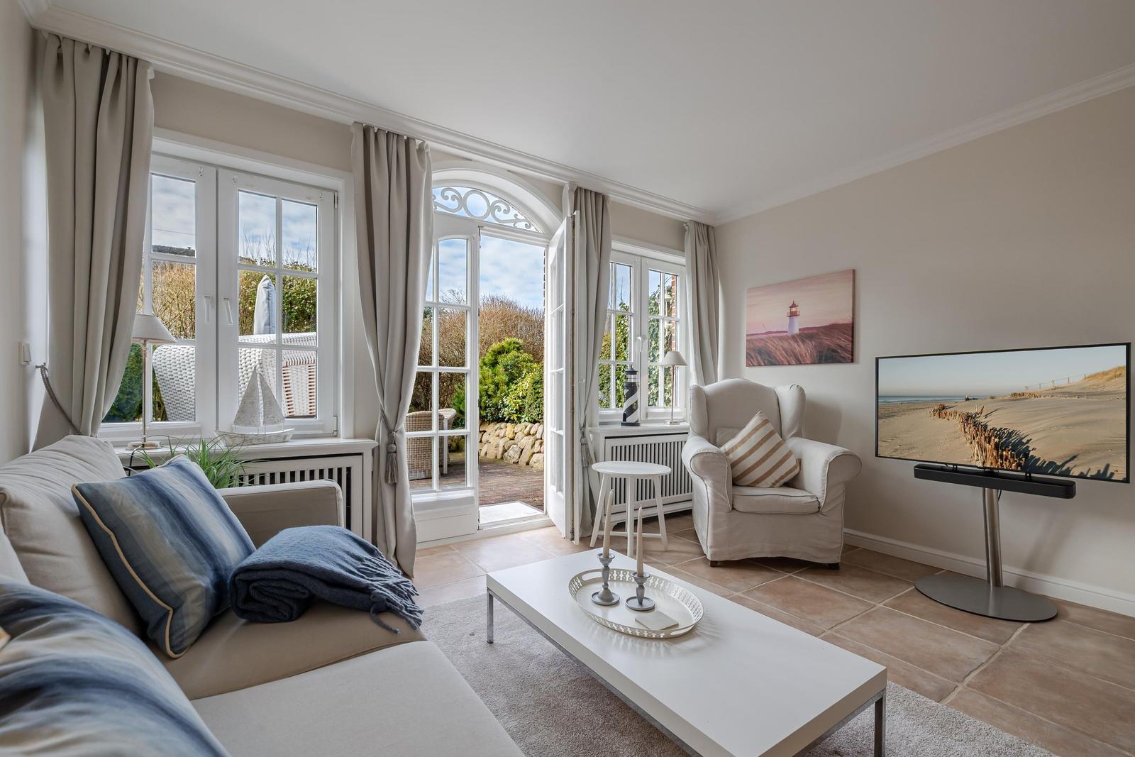 Wohnzimmer mit Sofa, Sessel und Fernseher. Durchgehende Fensterfront mit Blick auf Garten.