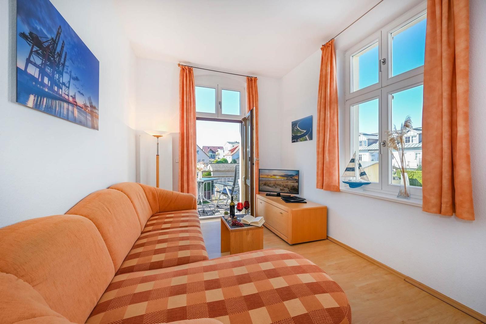 Wohnzimmer mit Sofa, TV und Balkon. Fenster mit orangefarbenen Vorhängen.