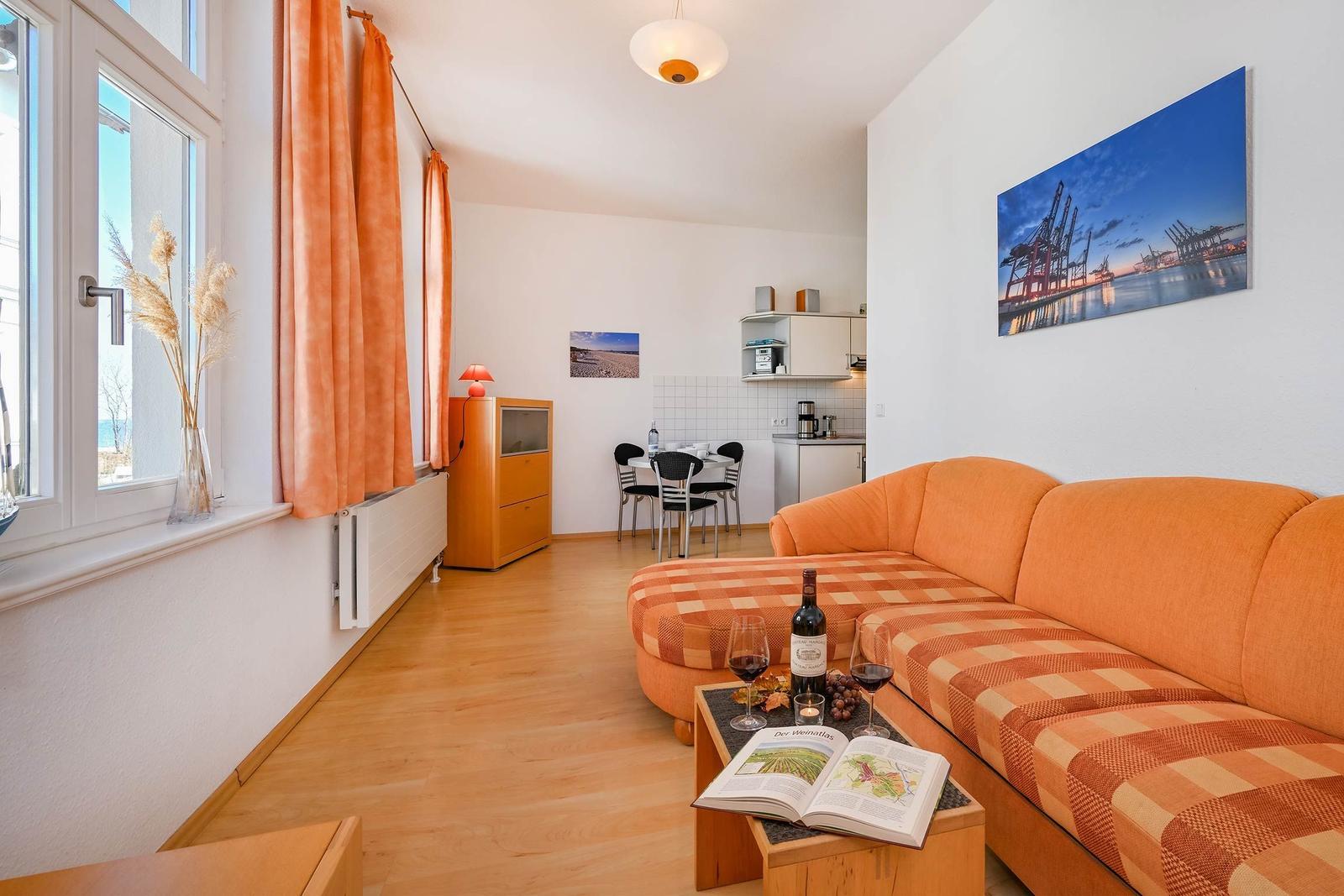 Wohnzimmer mit orangefarbenem Sofa, Couchtisch mit Wein und Buch, Fenster mit Vorhängen und Blick aufs Meer.