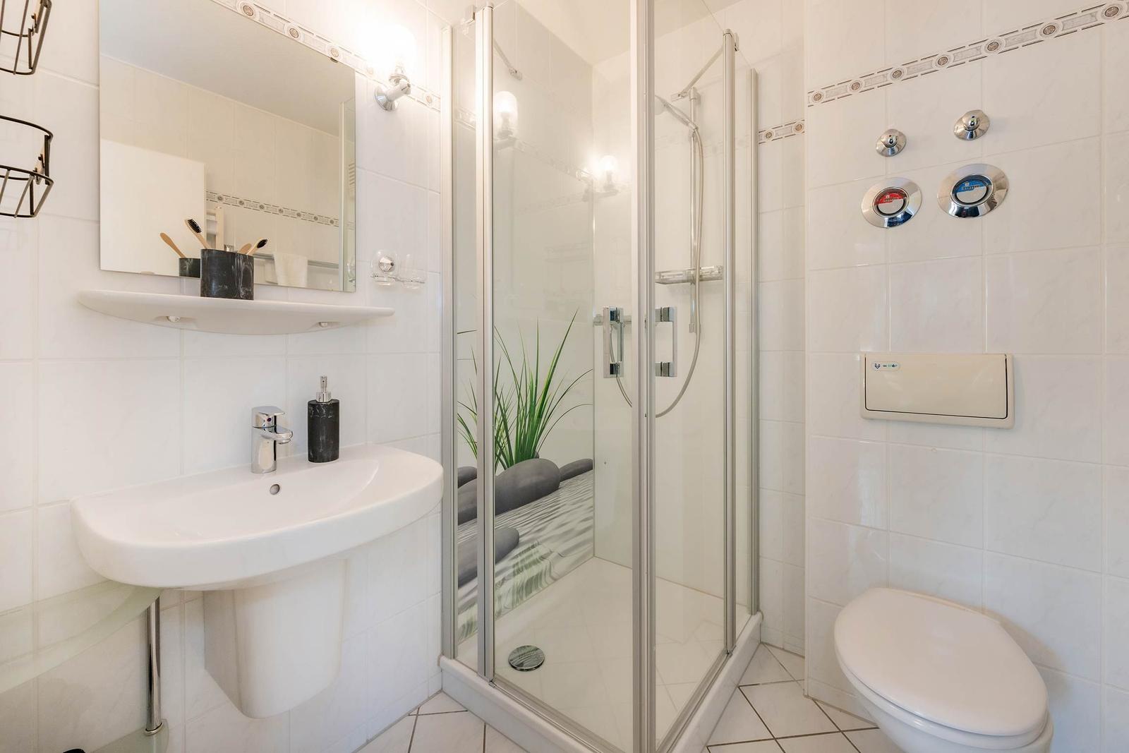Badezimmer mit Dusche, Waschbecken und Toilette. Weiße Fliesen und Spiegel.