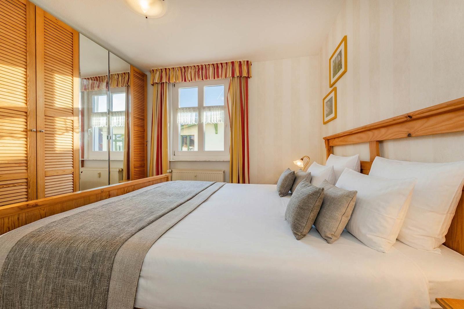 Schlafzimmer mit Holzbett, Spiegelschrank und Fenster mit Vorhängen.