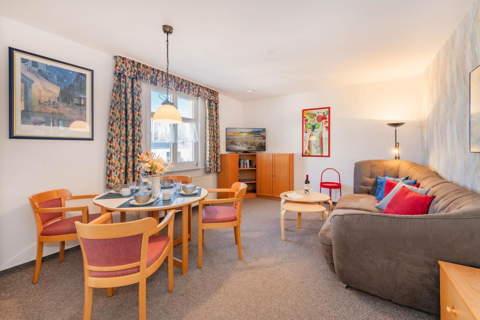 Wohn- und Essbereich mit Sofa, Tisch und Stühlen sowie Fenster und Wandbildern.