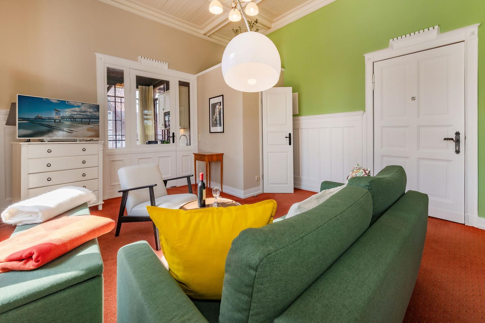 Ein Wohnzimmer mit grünem Sofa, gelben Kissen und rotem Teppich. Fernseher, Tür und Spiegel sind sichtbar.