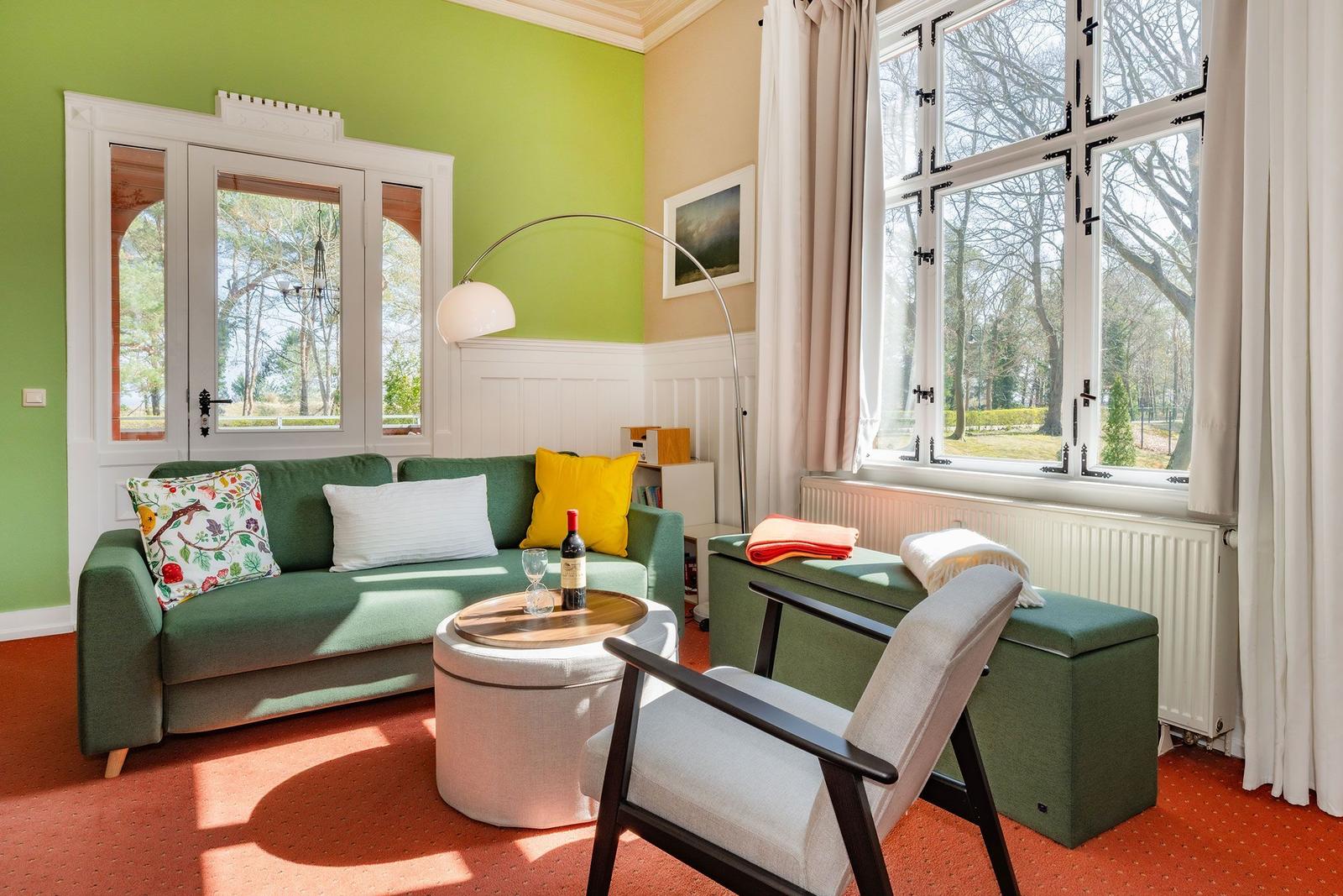 Ein Wohnzimmer mit grünem Sofa, runden Tisch und großen Fenstern.