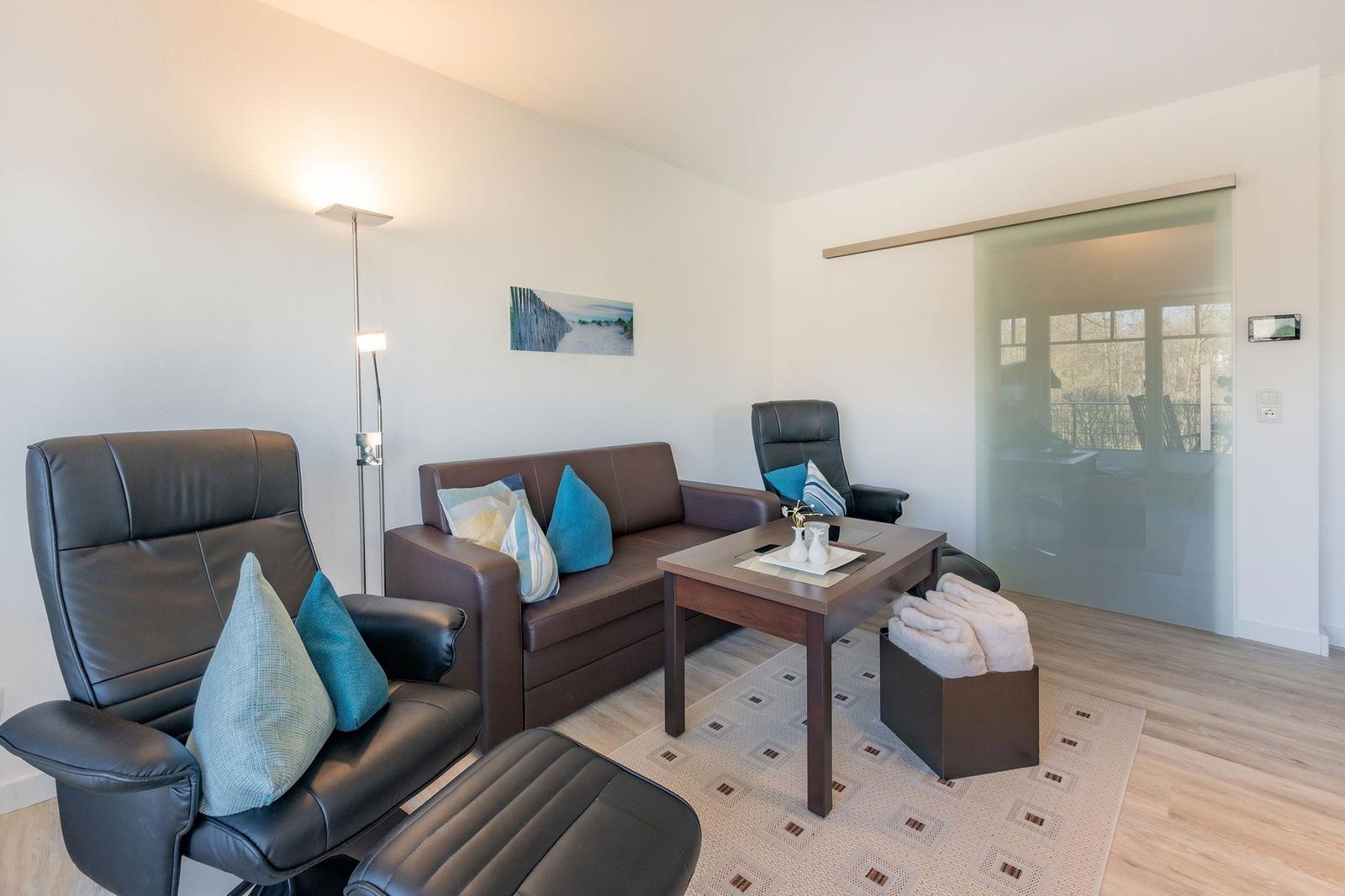 Living room with leather sofa, chairs and table. Entrance to terrace with glass wall.