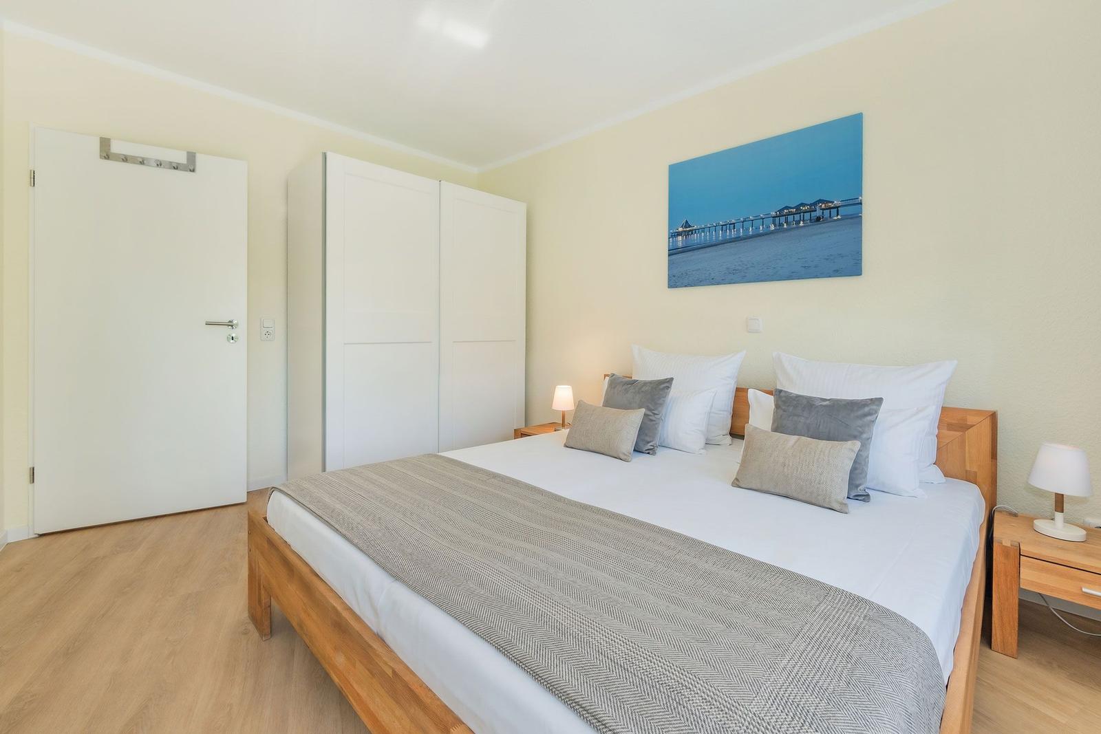 Schlafzimmer mit Bett, Nachttischen und Wandbild. Holzboden und weiße Möbel.