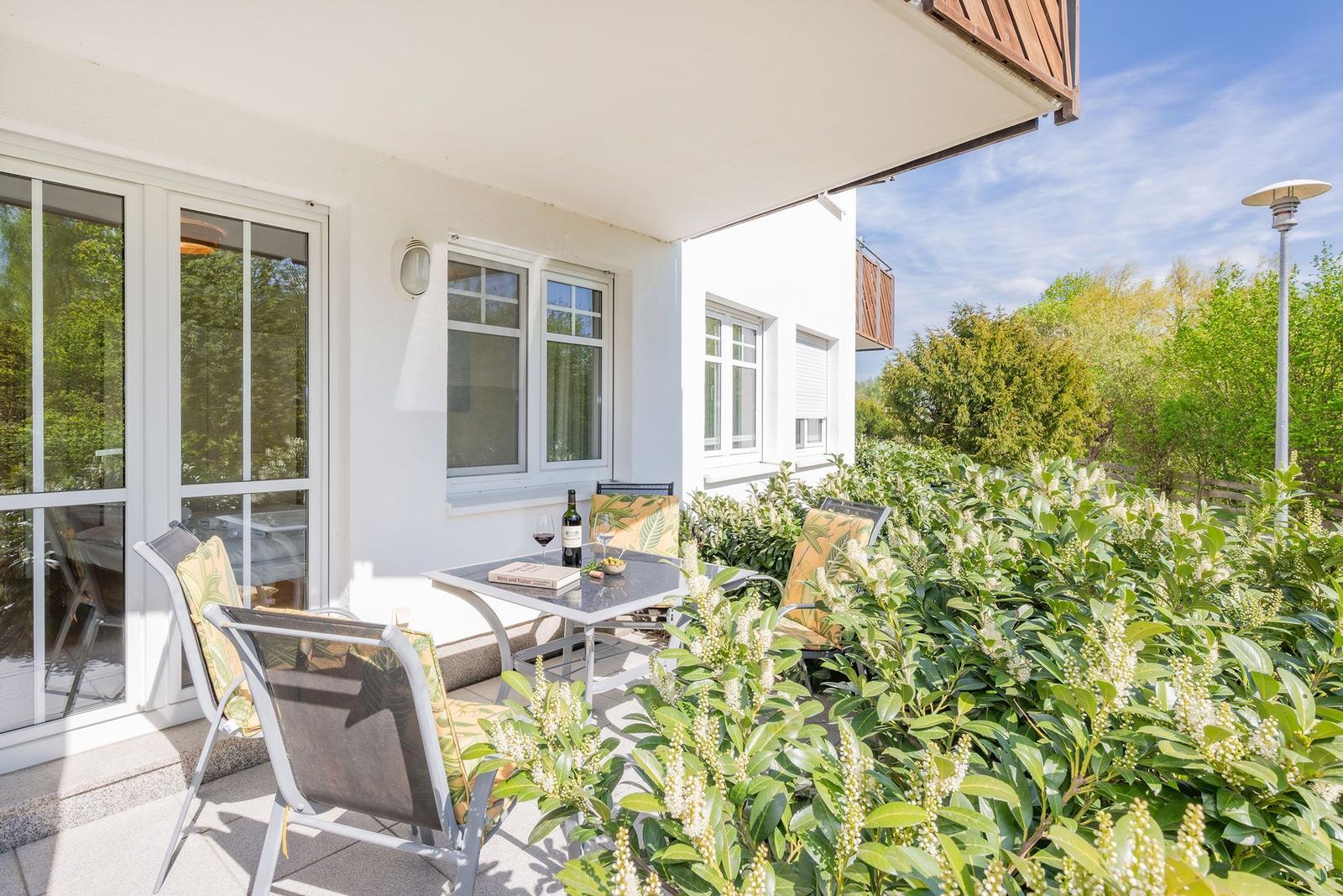 Terrasse mit Tisch, Stühlen und Weinflasche. Umgeben von grünen Pflanzen und blauem Himmel.