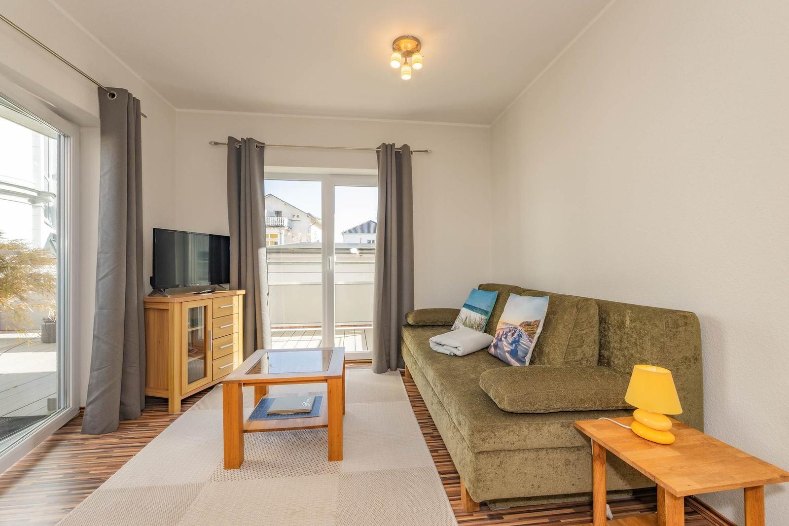 Wohnzimmer mit Sofa, Fernseher und Balkontür. Holzmöbel und Teppichboden.