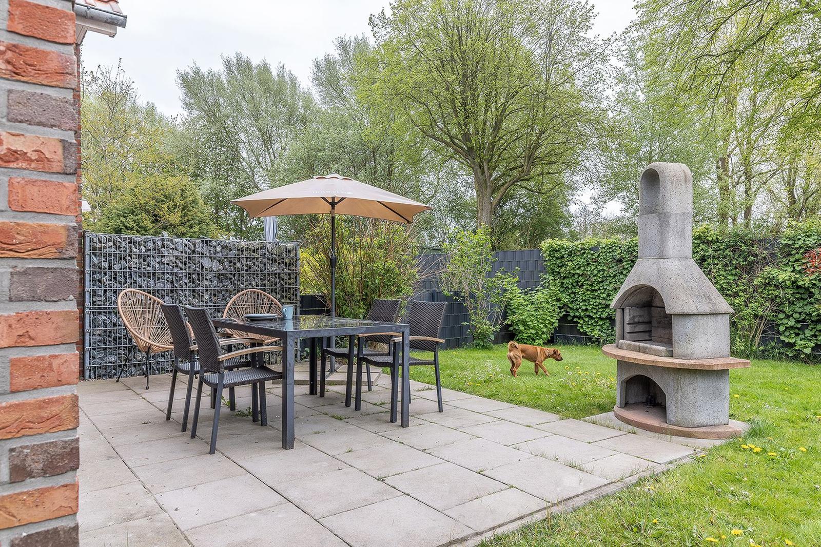 Terrace with table, chairs, and umbrella. Grill oven and lawn in the background.