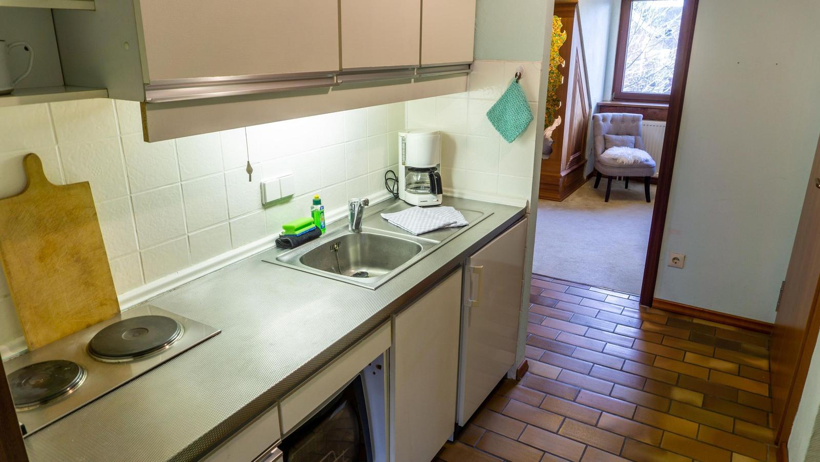 Küche mit Spüle, Kaffeemaschine und Kochfeld. Durchgang zu Wohnzimmer mit Sessel.