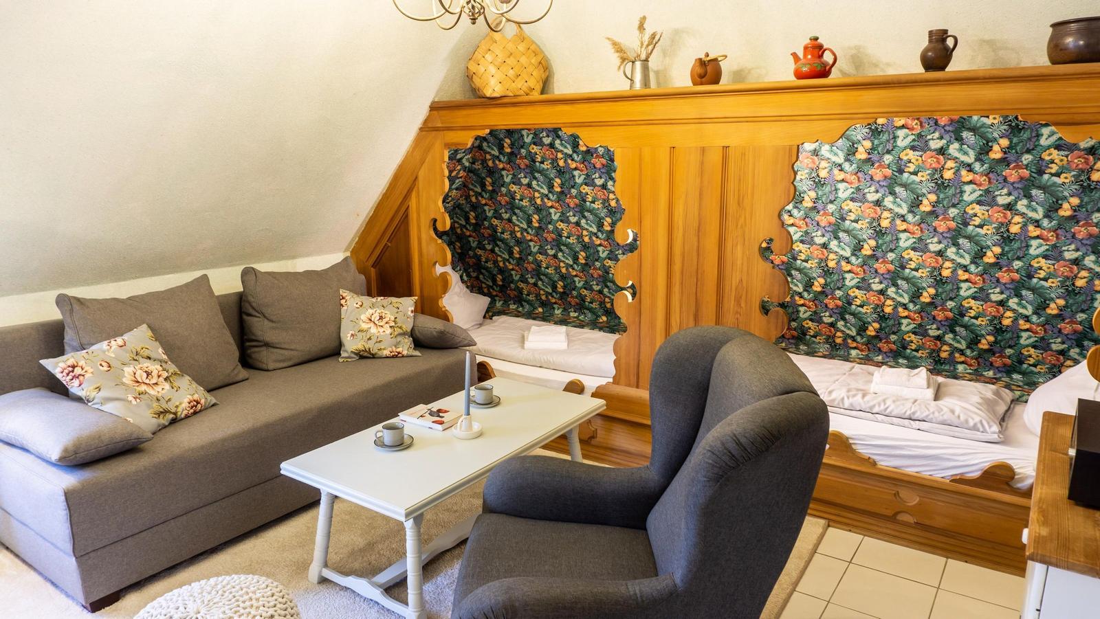 Zimmer mit Sofa, Sessel, Tisch und zwei Betten mit floraler Tapete.