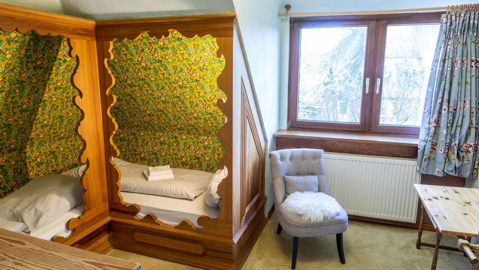 Zimmer mit Holzbett, Fenster, Stuhl und Tisch.