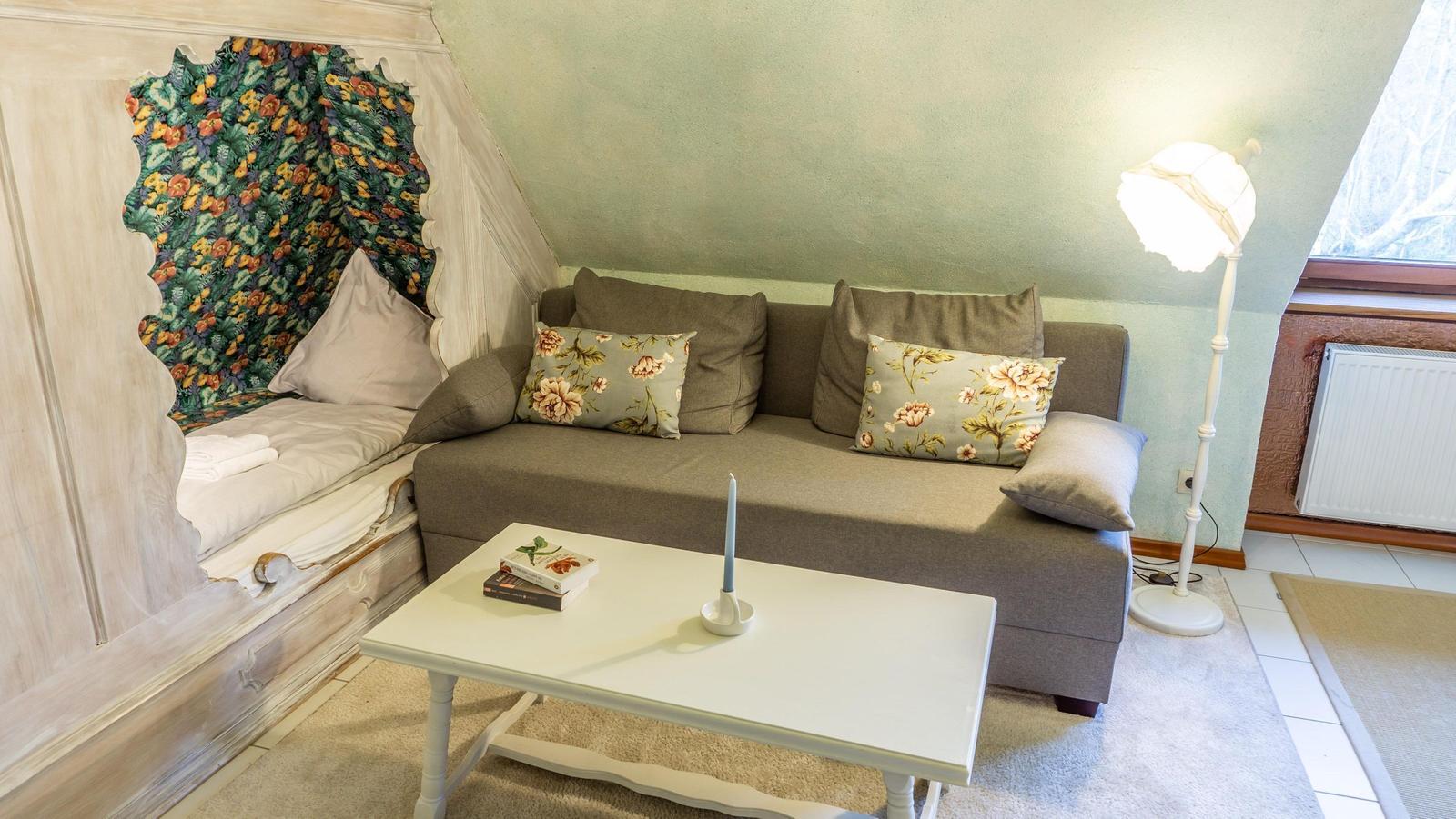 Ein Wohnzimmer mit Sofa, Couchtisch und Fenster. An der Wand ein Bett mit floraler Tapete.