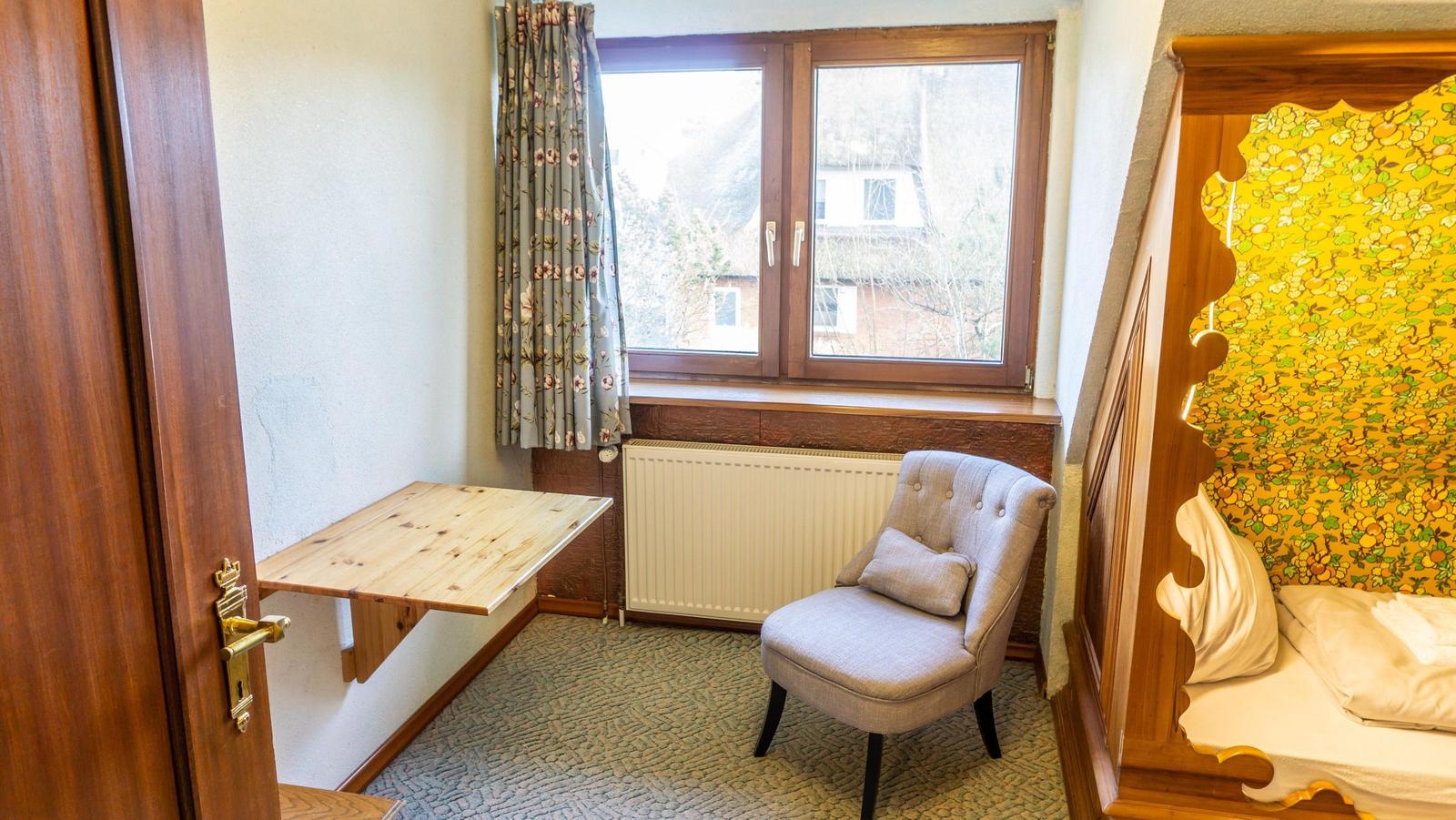 Ein Raum mit Schreibtisch, Sessel und Bett mit Holzrahmen und gelbem Tapetenmuster.