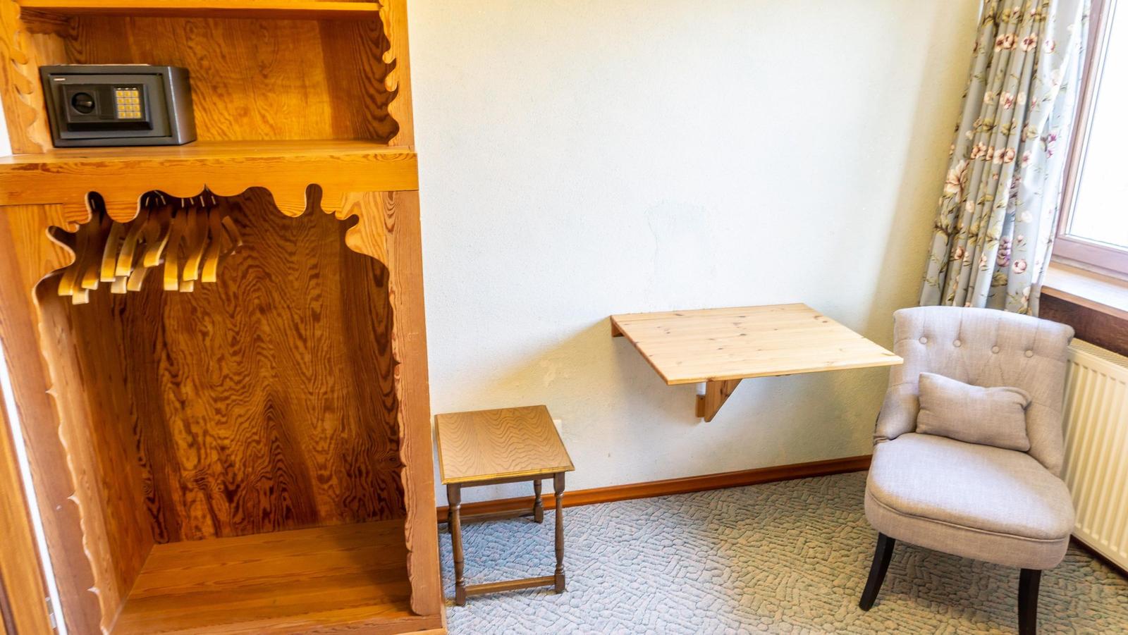 Ein Raum mit Kleiderschrank, Schreibtisch und Sessel. Vor dem Fenster hängen Vorhänge.