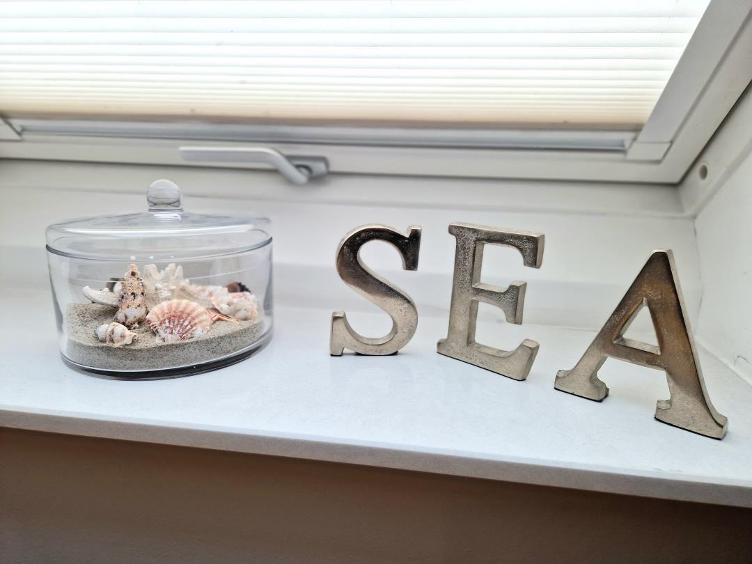 Auf der Fensterbank steht ein Glasbehälter mit Muscheln und Sand neben den Buchstaben SEA.