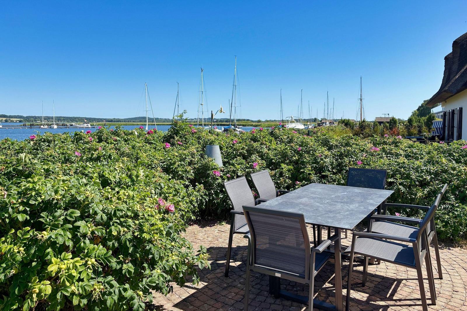 Terrasse mit Tisch und Stühlen, Blick auf Hafen mit Segelbooten und grüne Hecke.