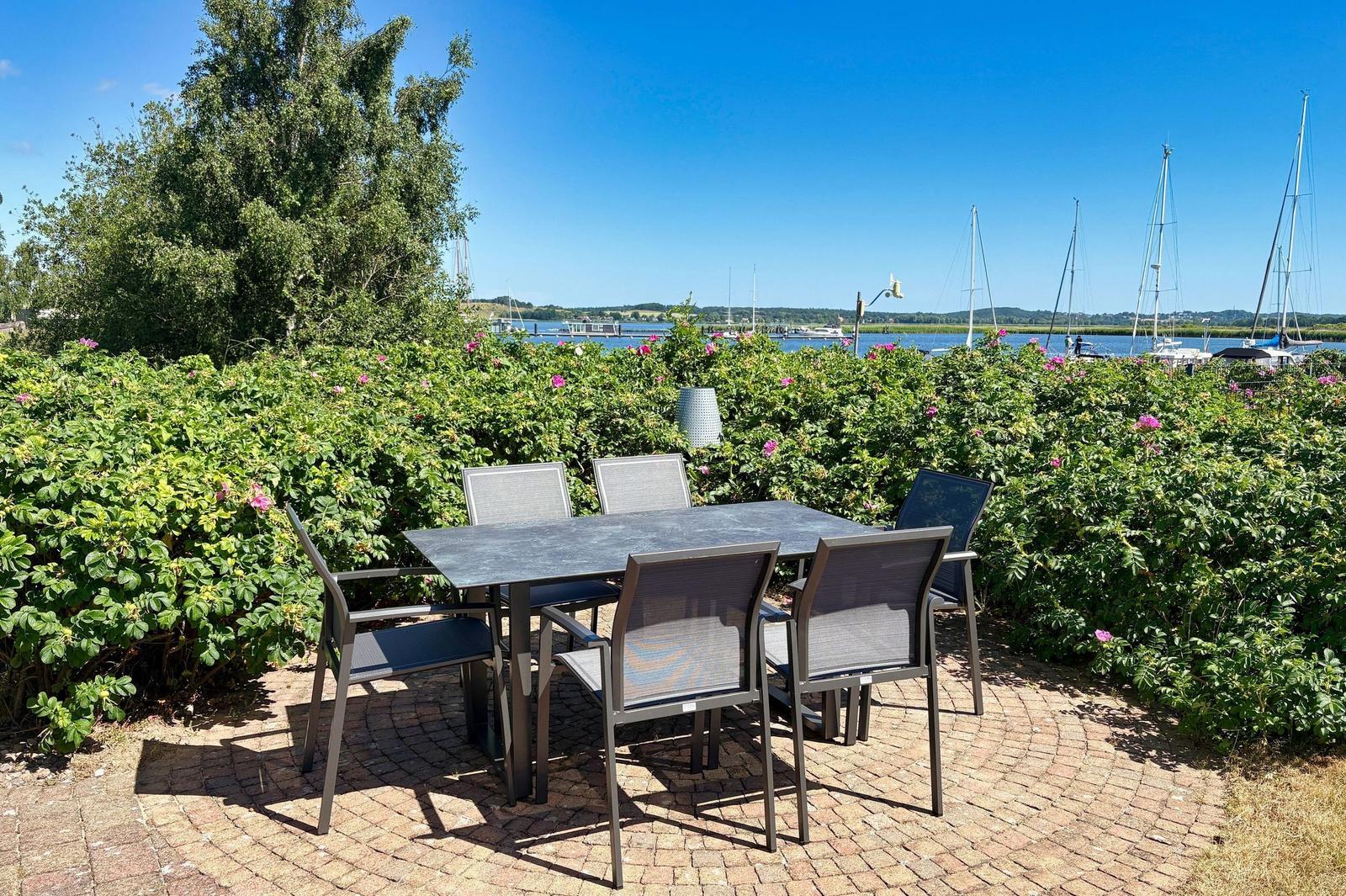 Terrasse mit Tisch und Stühlen, Blick auf Hafen mit Segelbooten und grüne Hecke.