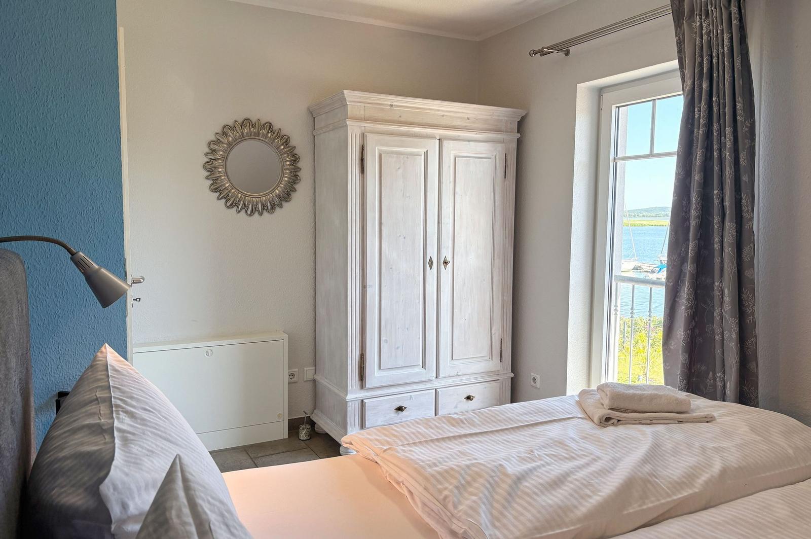 Schlafzimmer mit Bett, Kleiderschrank und Fensterblick auf Wasser.