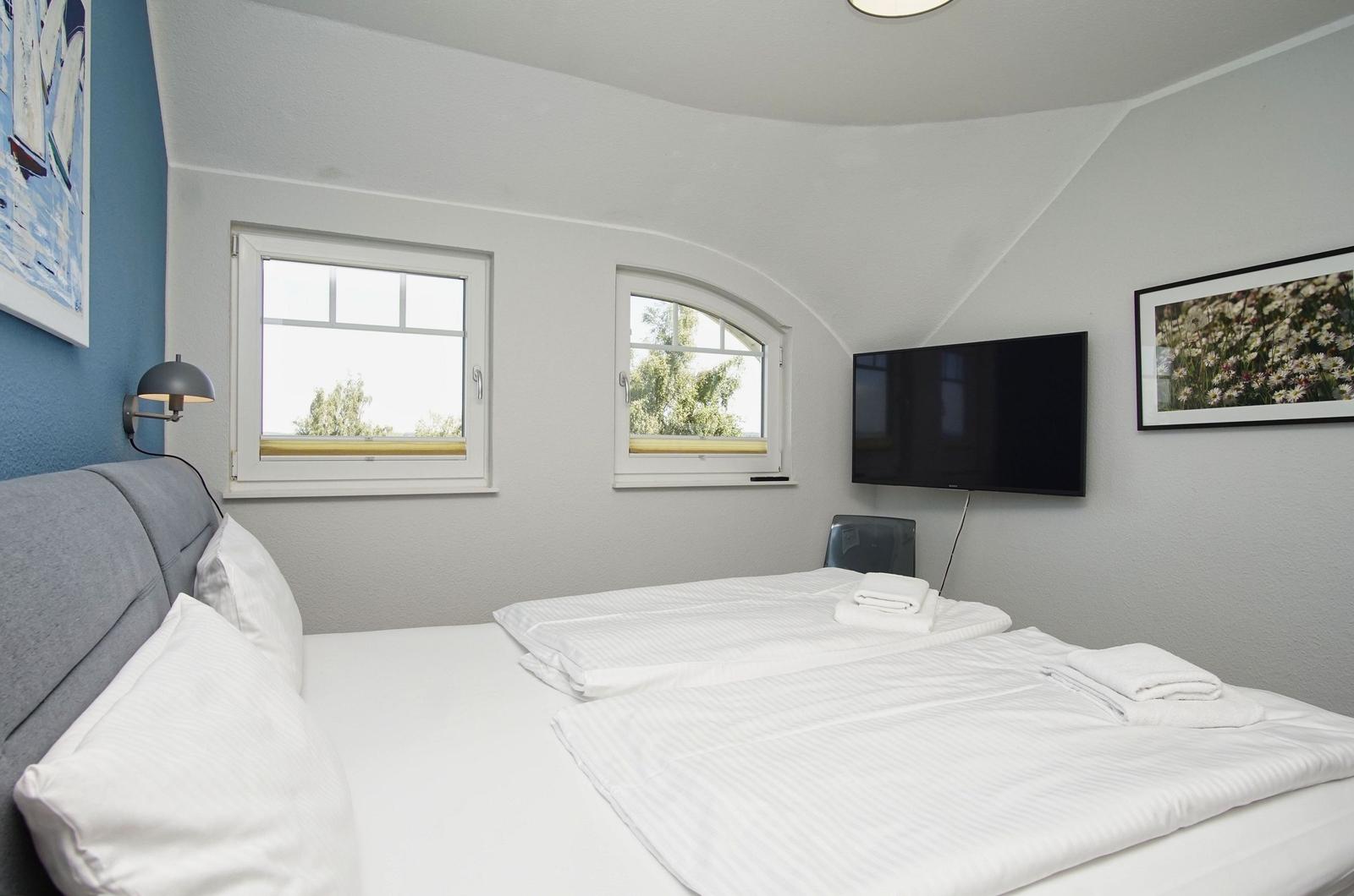 Schlafzimmer mit Doppelbett, zwei Fenstern und Wandfernseher.