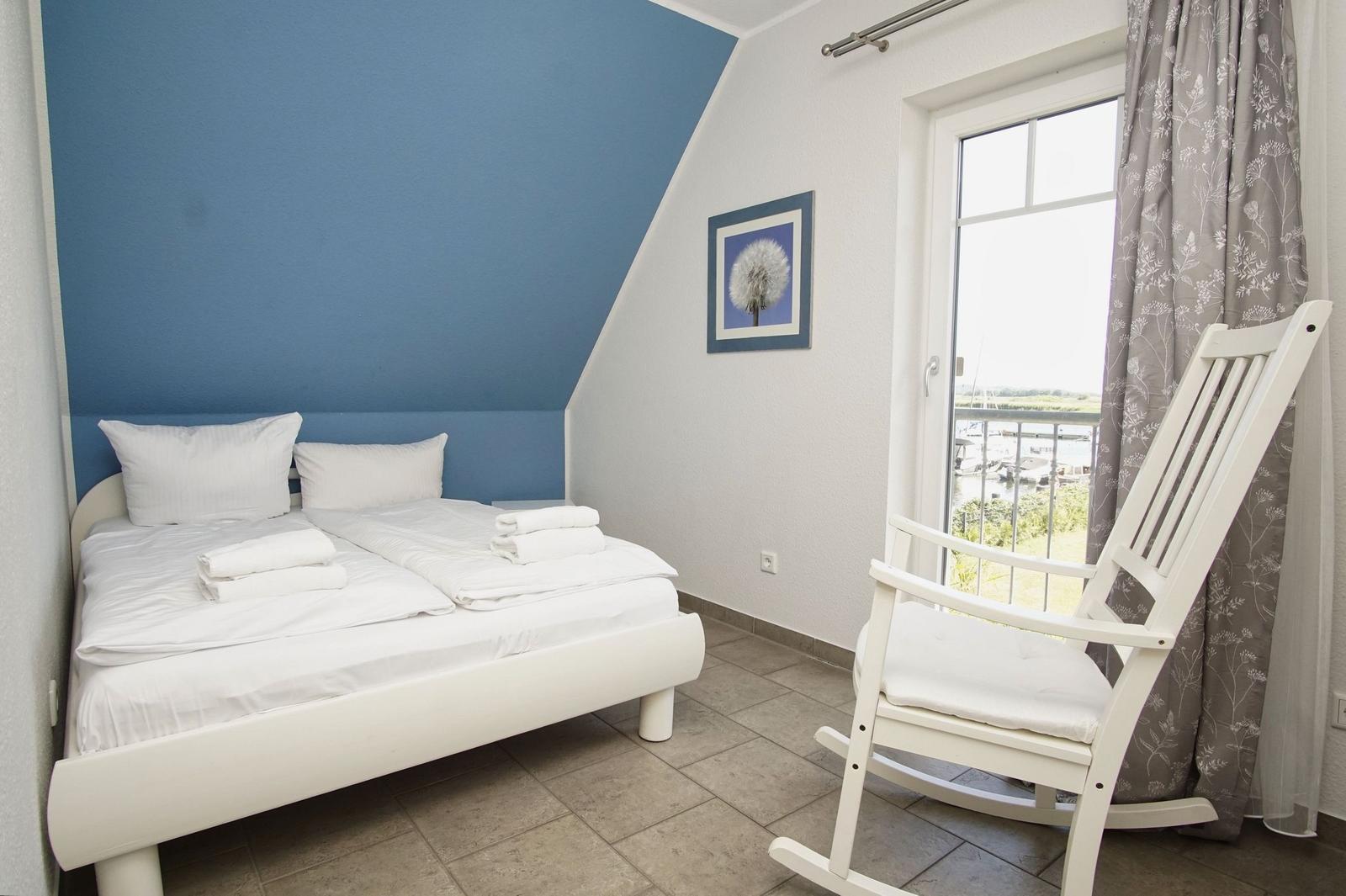 Schlafzimmer mit Doppelbett, weißem Schaukelstuhl und Blick auf den Hafen.