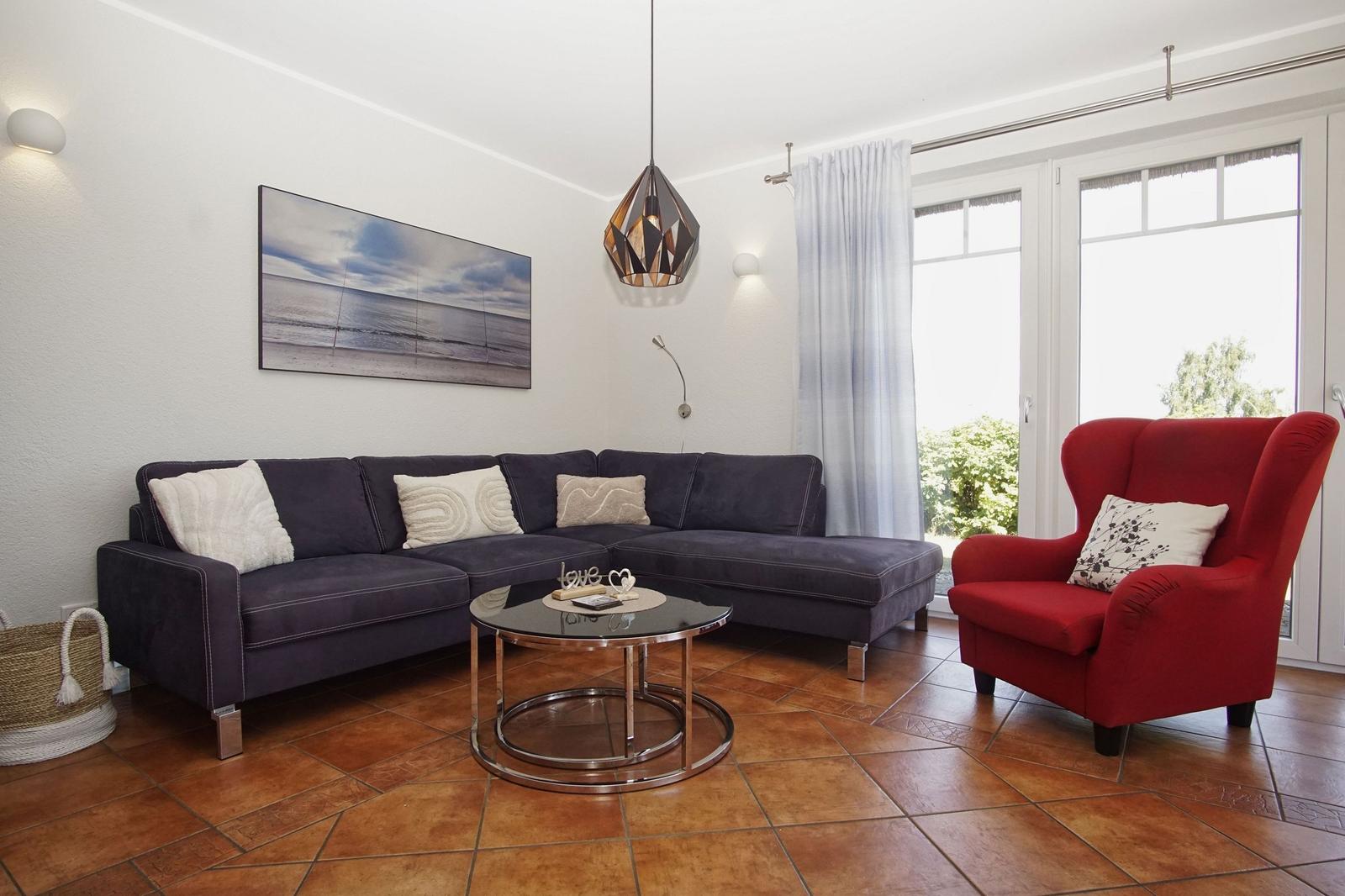 Ein Wohnzimmer mit dunkelblauem Sofa, rotem Sessel und Glas-Tisch. Fenster mit weißen Vorhängen.
