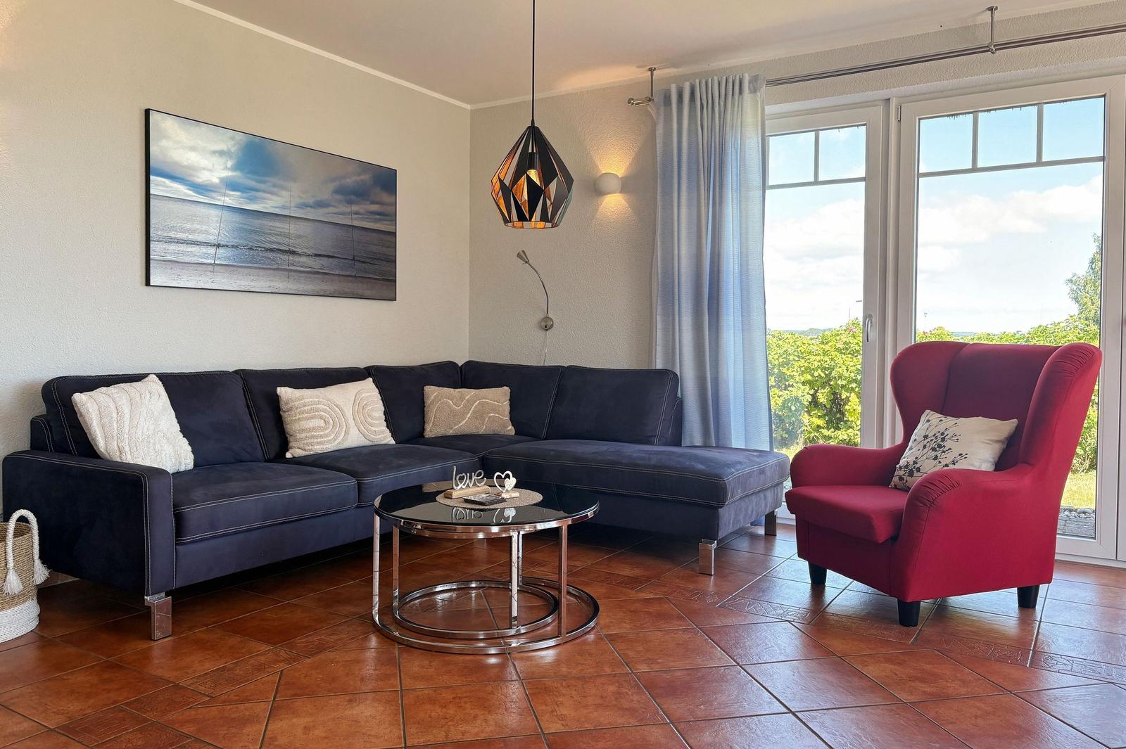 Ein Wohnzimmer mit dunkelblauem Sofa, rotem Sessel und Blick auf Grünflächen.