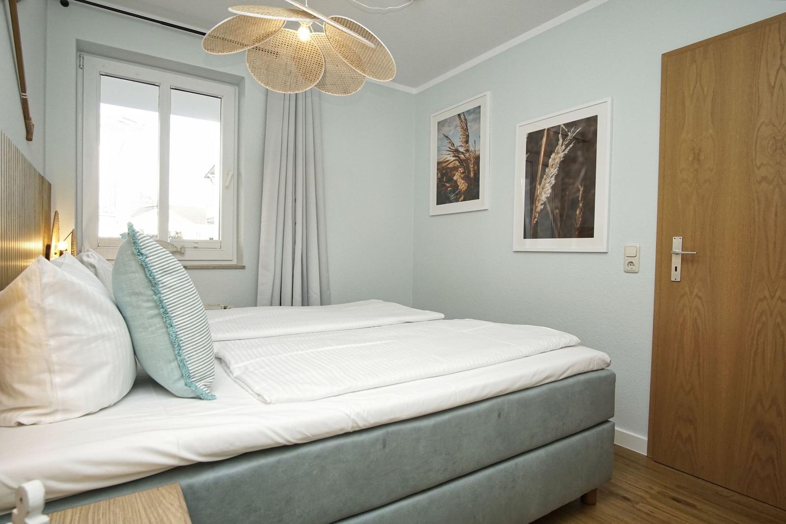 Schlafzimmer mit Bett, Fenster, Wandbildern und Deckenleuchte.