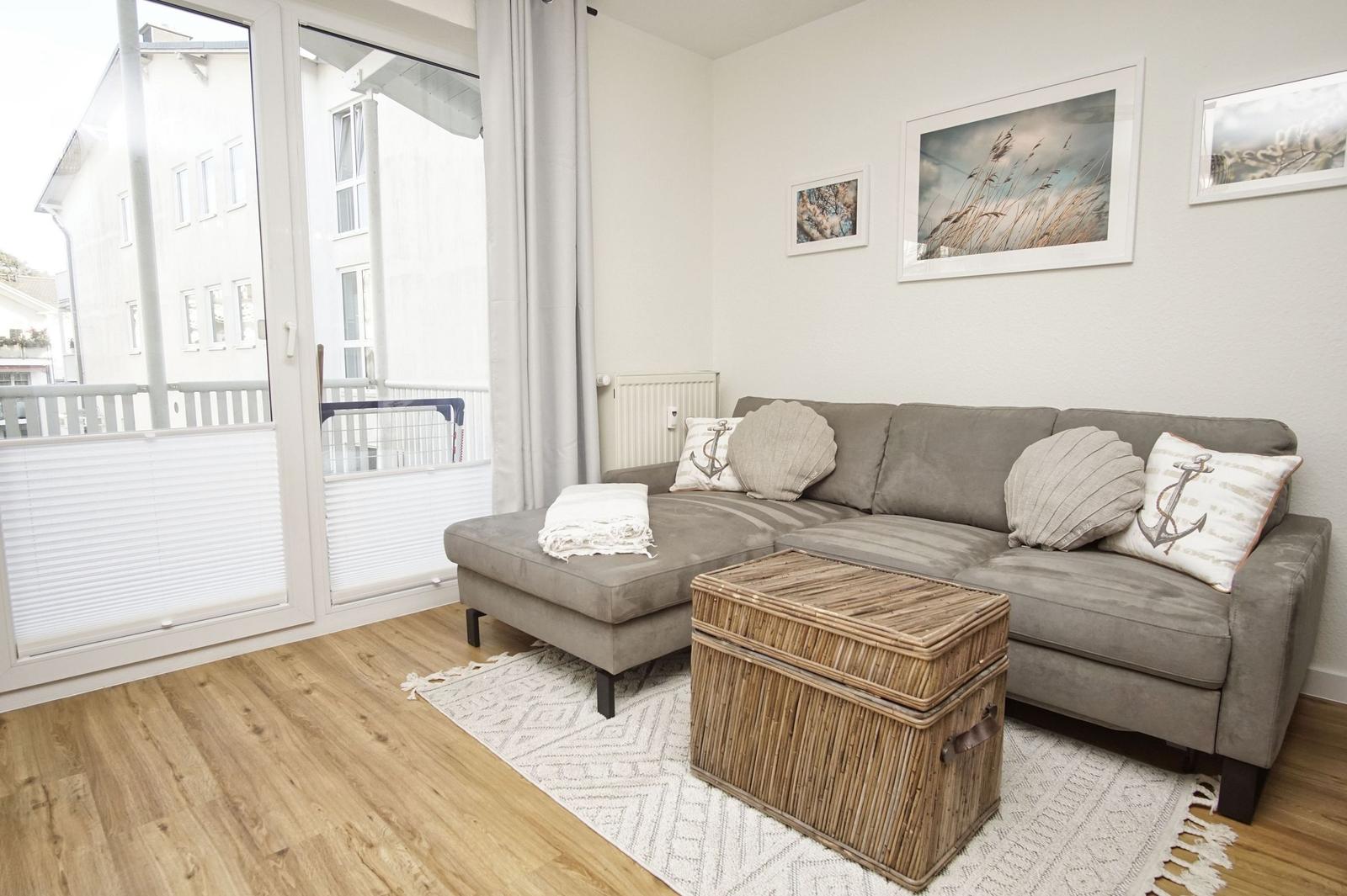 Ein Wohnzimmer mit grauem Sofa, Kissen und einem Korbtablett. Durchgehende Glastür zum Balkon.