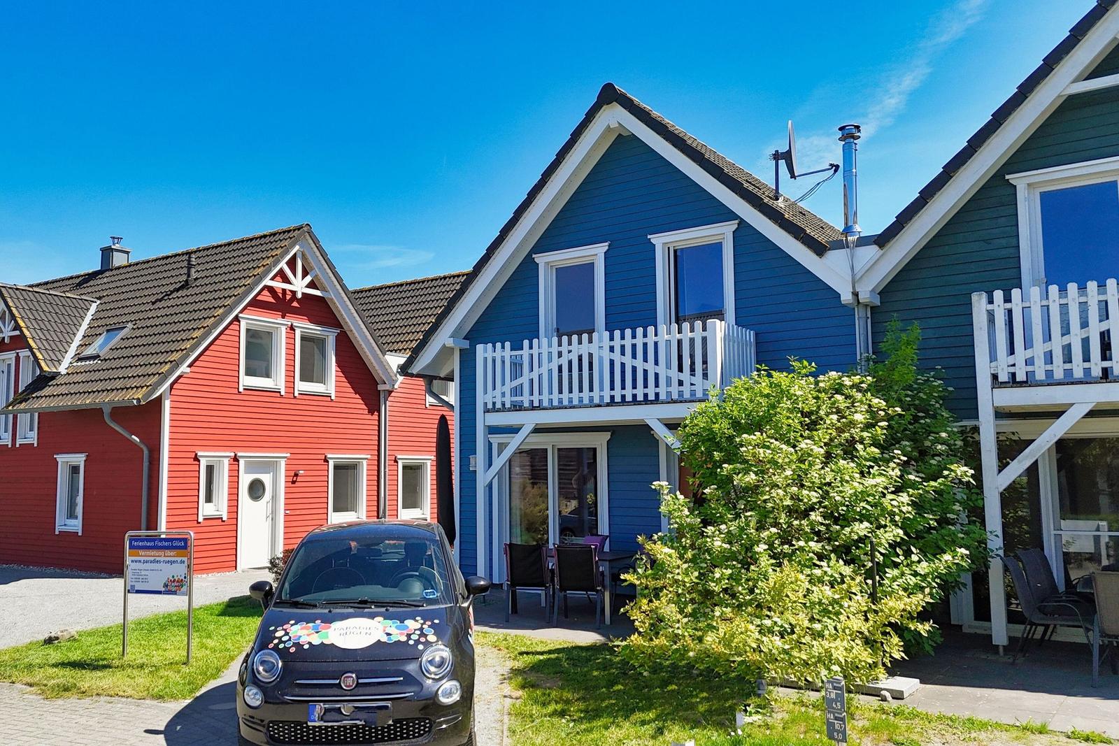 Blue vacation house with white balcony and garden area. A car is parked in front of the house.