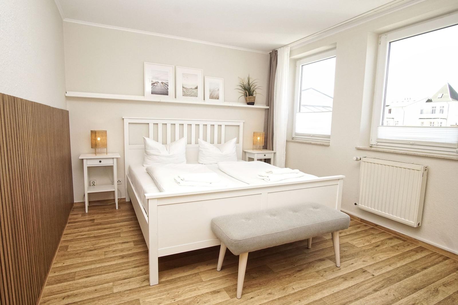 Schlafzimmer mit weißem Bett, Holzboden und Fenstern. An der Wand hängen Bilder.