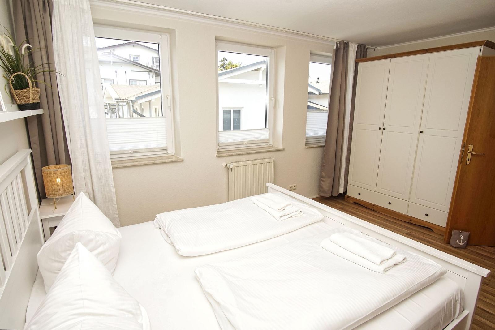 Schlafzimmer mit Doppelbett, Kleiderschrank und Fenstern mit Blick auf Gebäude.
