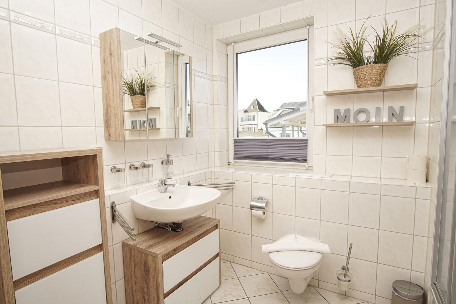 Badezimmer mit Waschbecken, Toilette und Fenster. Holzmöbel und Pflanze auf Regal.