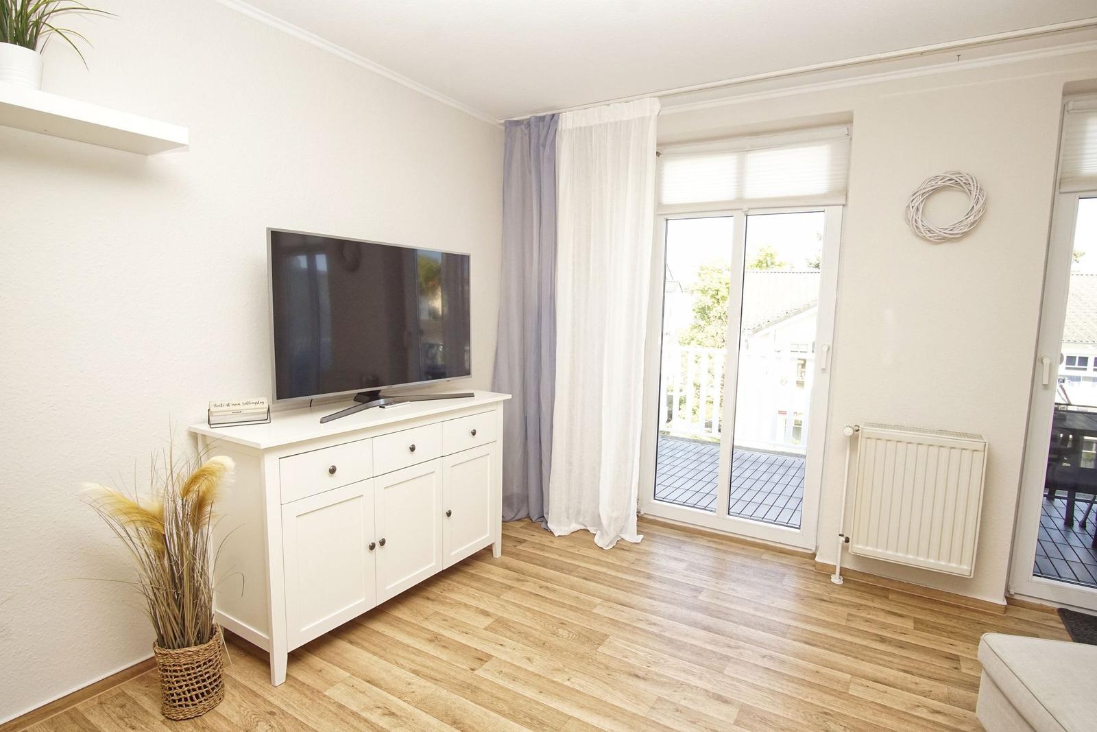 Wohnzimmer mit TV-Schrank, Holzboden und Balkontür. An der Wand hängt ein Kranz.