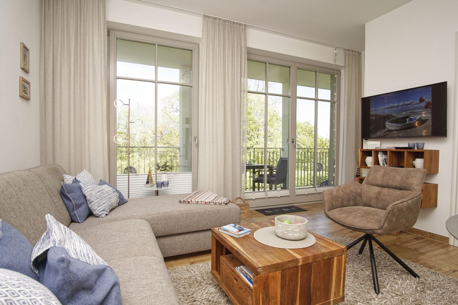 Wohnzimmer mit Sofa, Sessel, Couchtisch und TV. Große Fenster mit Blick auf Balkon.