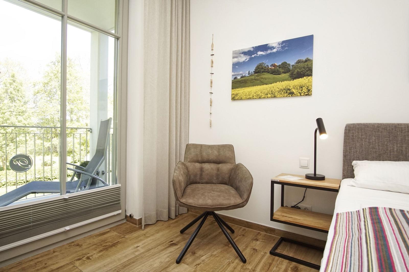 Zimmer mit Bett, Stuhl und Balkonansicht. Wandbild und Nachttisch mit Lampe.