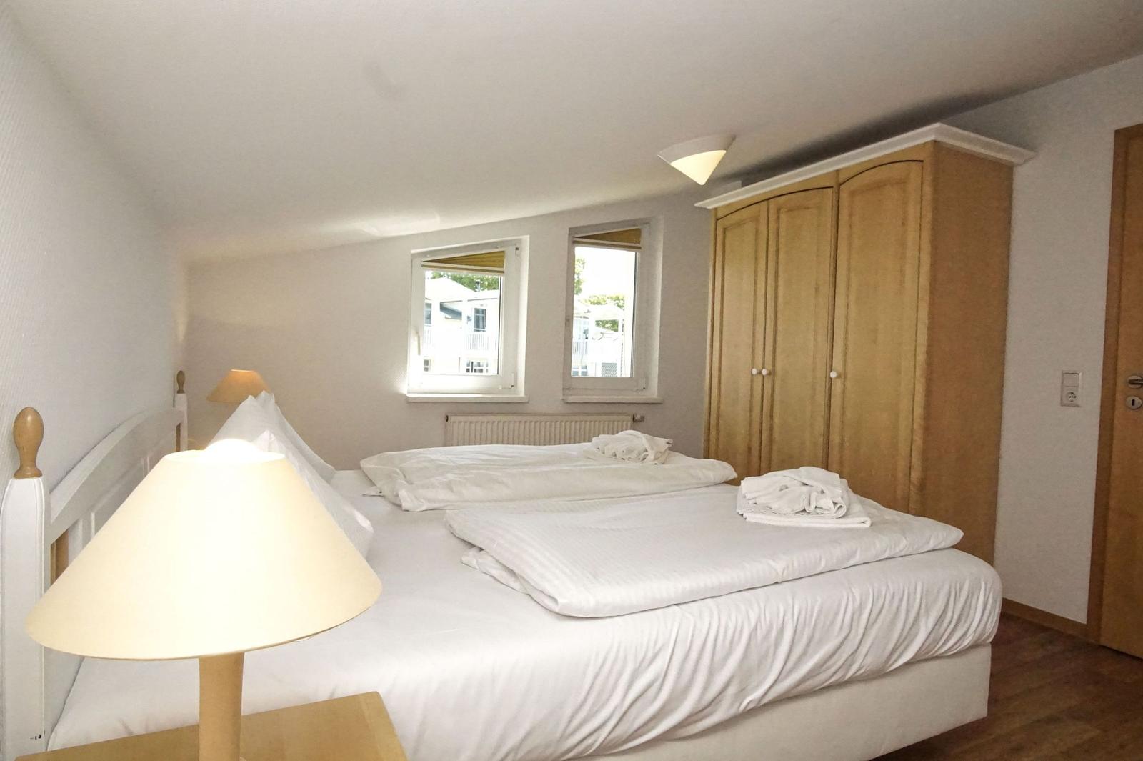 Schlafzimmer mit Doppelbett, Holzschrank und zwei Fenstern.