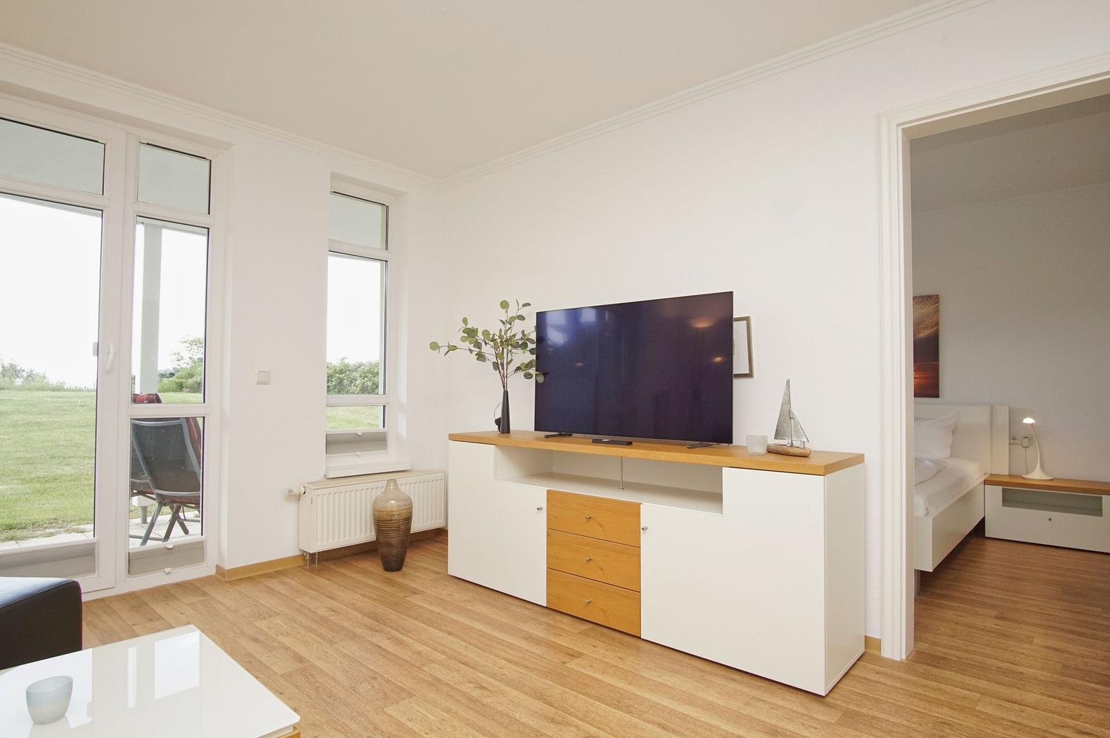 Living room with TV unit, windows, and view of green area. Bedroom visible in background.