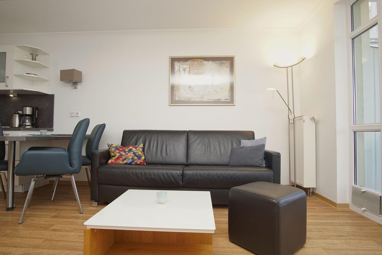 Living and dining area with sofa, table and chairs. Windows and wall lamps visible.