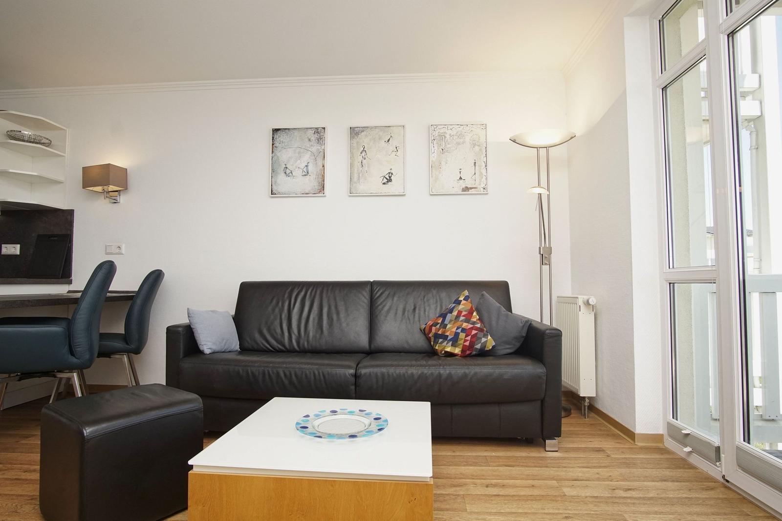 Wohnzimmer mit Sofa, Couchtisch und großen Fenstern. Wandbilder und Stehlampe sichtbar.